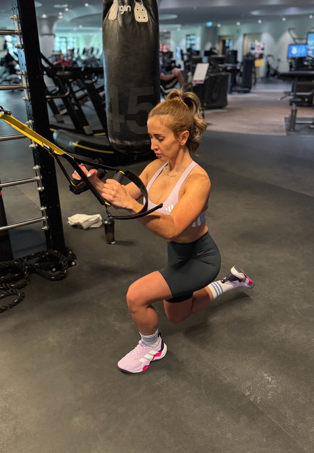 Mujer realizando el ejercicio de zancada con bandas de suspensión en un gimnasio, con un saco de boxeo y equipo de fitness de fondo.