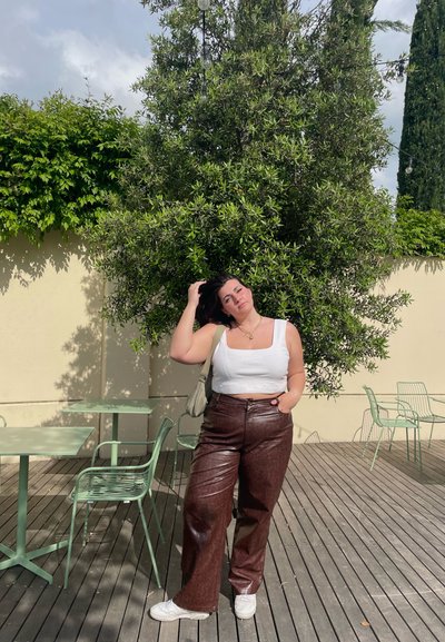 Top corto blanco, pantalones anchos de imitación cuero marrón con textura estampada, zapatillas blancas, de pie junto a mesas de café de metal verde.