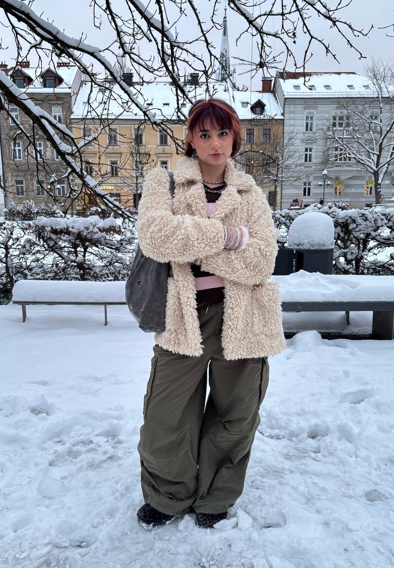 Manteau crème duveteux à manches longues porté sur un haut superposé rose et noir ; pantalon cargo ample vert olive ; sac gris ; arrière-plan enneigé.