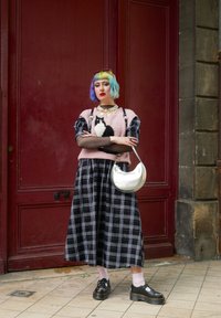 Το πουλόβερ διαθέτει ροζ πλέξιμο με σχέδιο μαύρης γάτας, συνδυασμένο με μια μακριά φούστα σε μαύρο και λευκό καρό. Ασημένια τσάντα με μορφή ημισελήνου και μαύρες πλατφόρμες.