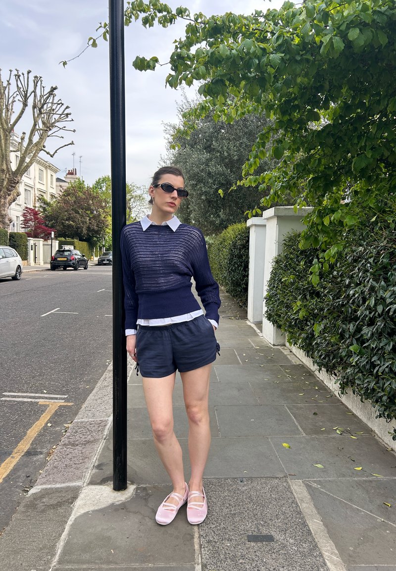 Femme portant des lunettes de soleil, un pull bleu marine, une chemise blanche à col et un short bleu marine s'appuie sur un poteau noir sur un trottoir avec de la verdure et des maisons à proximité.