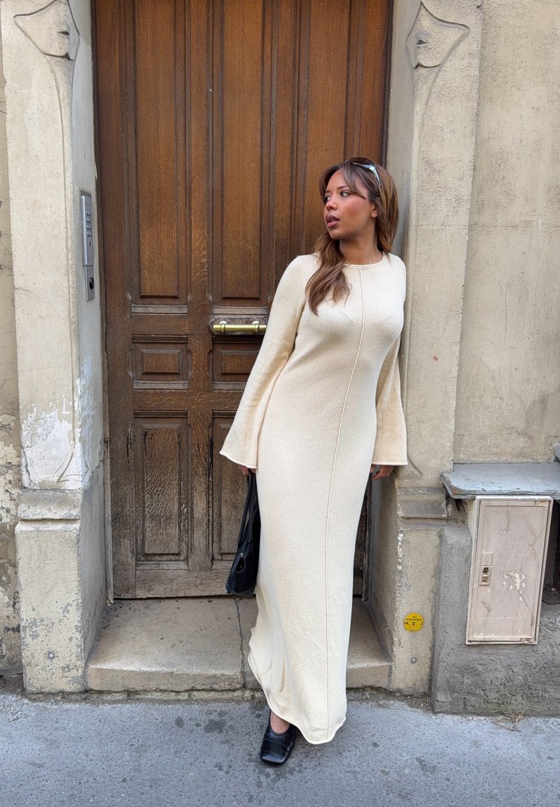 Femme en longue robe crème et chaussures noires tenant un sac noir, debout près d'une porte en bois encadrée dans une façade en pierre usée.