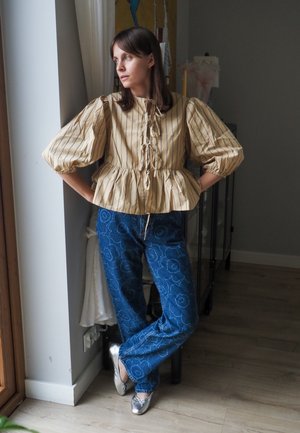 Blusa amarilla a rayas con mangas abullonadas, cintura ceñida y parte frontal con cordones; jeans de denim azul con estampado; zapatos planos plateados.