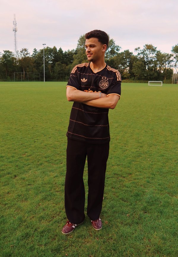 Jeune homme en maillot et pantalon de football noirs, bras croisés, debout sur un terrain de football herbeux au crépuscule.