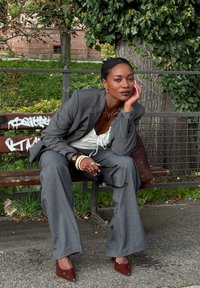 Grau Anzug mit weit geschnittenen Hosen und tailliertem Blazer. Weiße Bluse darunter, geschichteter Schmuck und kastanienbraune High Heels. Sitzt auf einer Bank.