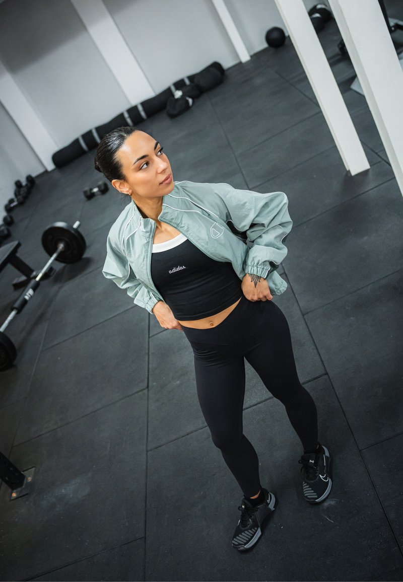 Sportieve outfit met een lichtgroene bomberjack, een zwarte crop top met het "adidas"-logo en zwarte legging. Zwarte sportschoenen met een textuurontwerp.