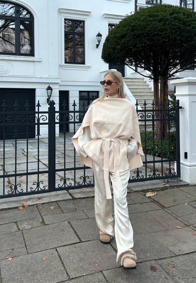 Poncho de lino beige con cintura ceñida con cinturón, combinado con pantalones crema brillantes. Accesorios incluyen gafas de sol negras y calzado beige forrado de piel.