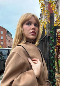 Manteau en laine marron clair avec des manches larges, doté d'une ceinture, associé à un sac à bandoulière noir. Texture douce, décor extérieur avec du feuillage d'automne.