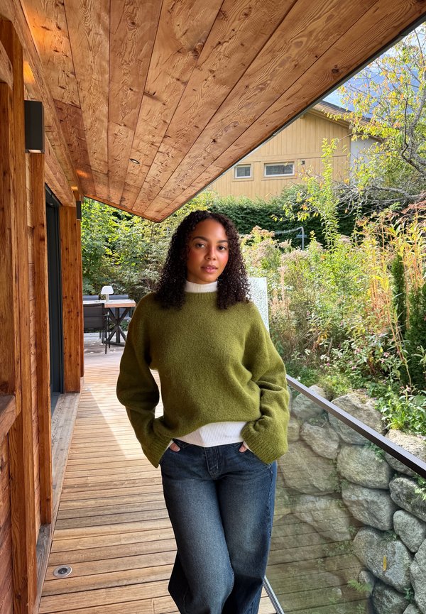 Une femme portant un pull vert et un jean se tient sur une terrasse en bois, près d’une balustrade en verre, entourée de verdure.