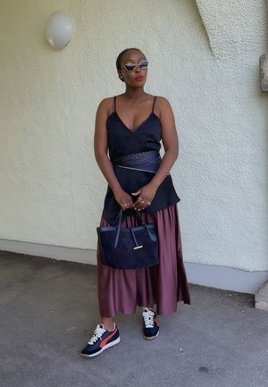 Top de satén negro, falda fluida burdeos, zapatillas de deporte azul marino con acentos naranjas. Bolso de ante negro. Diseño minimalista, gafas de sol elegantes.