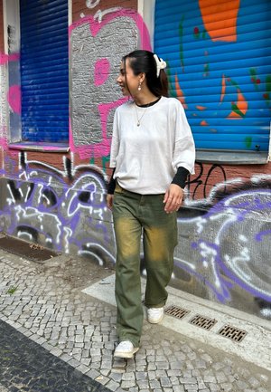 Camiseta de gran tamaño gris claro con mangas negras, pantalones verdes con un tono amarillo y zapatillas blancas. Pelo recogido con una goma blanca.