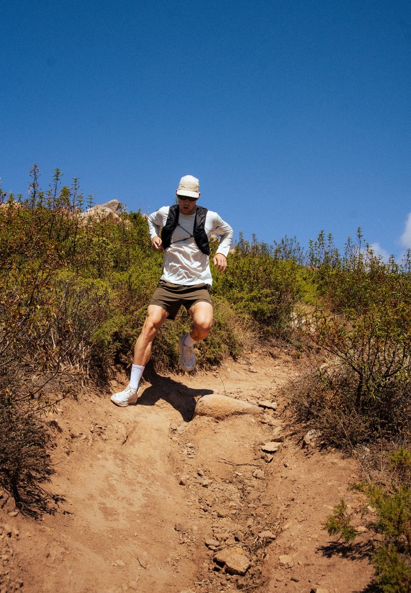 Coureur en débardeur noir et chemise à manches longues blanche, portant un short beige et des baskets blanches, naviguant sur un sentier de terre rocailleux entouré de verdure.