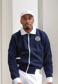 Giacca zip-up blu navy con colletto e dettagli bianchi, caratterizzata da una toppa con logo. La persona indossa un cappello bianco e pantaloni bianchi.