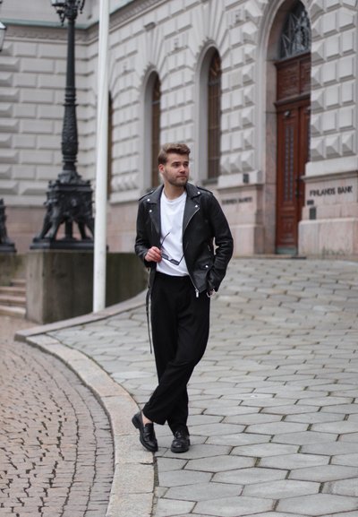 Chaqueta de cuero negro de motorista sobre una camiseta blanca, combinada con pantalones negros holgados y zapatos negros. El modelo está de pie en unos escalones de adoquines.