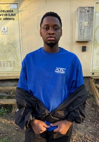 Blauw T-shirt met een wit logo op de linkerborst, gecombineerd met een zwart jack met rits. De scène toont een beige muur en een elektriciteitskast.