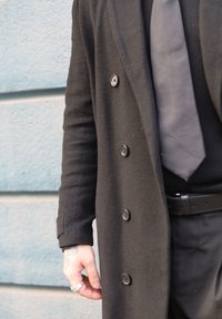 Manteau noir croisé en matière texturée, avec des boutons proéminents et une coupe slim. Main visible portant une bague, sur fond bleu.