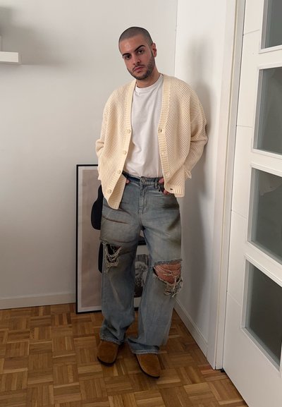 Cardigan de punto crema sobre una camiseta blanca, combinado con jeans desgarbados azul claro con rotos en las rodillas y zapatos marrones tipo slip-on.