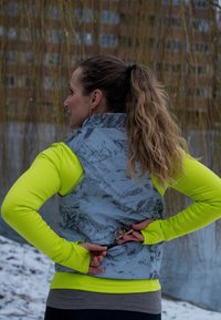 Femme avec queue de cheval portant une tenue de sport jaune fluo et grise ajuste une petite poche zippée sur son gilet en plein air près du sol enneigé et de l'eau.