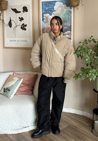 Chaleco de plumas beige con cierre de cremallera y mangas oversized, combinado con pantalones holgados negros y botas negras voluminosas, contra un fondo neutro.