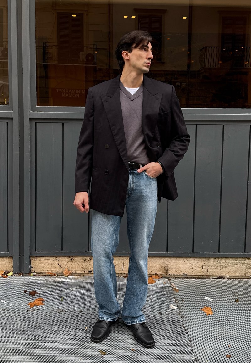 Homme debout sur un trottoir urbain portant un blazer noir, un pull gris, un jean bleu clair et des chaussures noires, regardant sur le côté avec la main dans la poche.