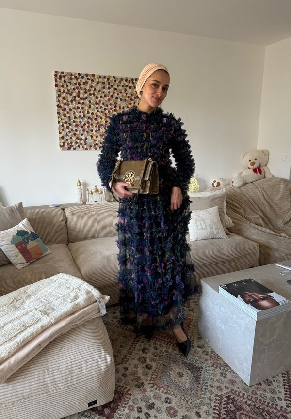 Femme portant une robe florale texturée foncée et un foulard beige, tenant un sac à main marron, debout dans un salon avec un canapé beige et des coussins décoratifs.