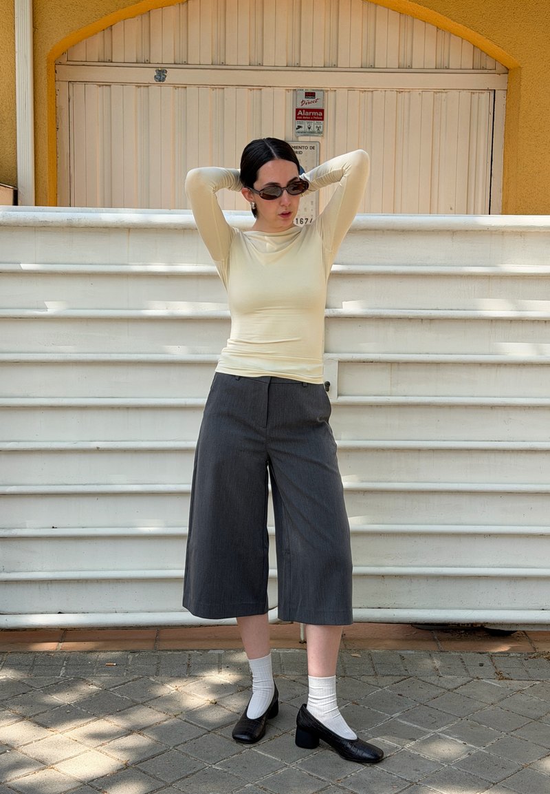 Femme portant des lunettes de soleil, un haut à manches longues crème, un pantalon large gris, des chaussettes blanches et des chaussures noires, debout avec les mains derrière la tête.