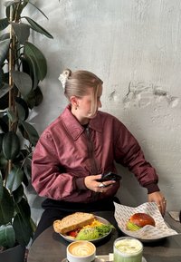 Bordo bomber jakna, smešna kosa u punđi, drži telefon. Stol s višebojnom salatom, sendvičem, burgerom i zelenim napitkom.