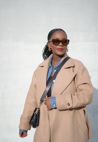 Ανοιχτό καφέ μάλλινο παλτό, τζιν πουκάμισο με ρίγες, μαύρη τσάντα χειρός με χρυσές λεπτομέρειες, υπερμεγέθεις γυαλιά ηλίου και σκουλαρίκια κρίκοι.