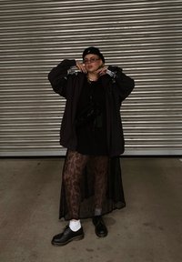 Μαύρο υπερμεγέθες μπουφάν με λευκές ρίγες, μαύρη μπλούζα, διαφανής δαντελένια φούστα και μαύρα παπούτσια με χοντρές σόλες. Τα αξεσουάρ περιλαμβάνουν γυαλιά ηλίου και τσάντα.