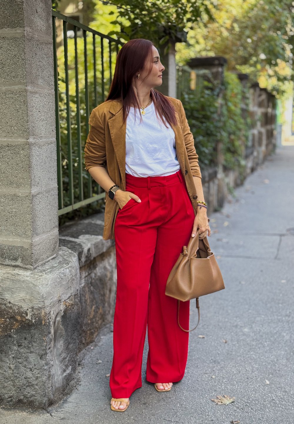 Sacou din suede maro, tricou alb, pantaloni largi roșii. Geantă maro, sandale. Gard verde și zid de piatră în fundal. Cadru urban casual.
