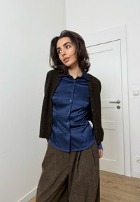 Szatén tengerészkék blúz gombos elől, texturált sötétzöld kardigánnal párosítva. A modell barna, bőszárú nadrágot visel. Fafelületű padló és fehér ajtó látható.