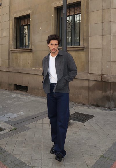 Chaqueta de mezcla de lana gris, camiseta blanca, vaqueros de mezclilla azul oscuro, zapatos negros; de pie contra una pared de piedra con ventanas al fondo.