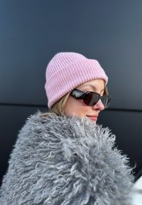 Femme portant un bonnet tricoté rose, des lunettes de soleil Prada noires et un manteau gris duveteux sur un fond sombre uni.