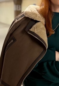 Veste en shearling marron avec doublure en fourrure beige et fermetures éclair argentées. Gros plan sur la zone de l'épaule, mettant en valeur les détails du matériau et des coutures.