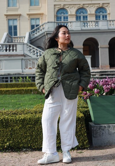 Joven mujer con cabello largo y oscuro lleva chaqueta acolchada verde y pantalones blancos, de pie en un jardín con flores y un edificio histórico de fondo.