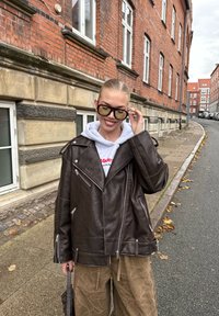 Jachetă de motociclist din piele maro cu fermoare argintii, purtată peste un hanorac gri. Persoana ține ochelari de soare negri, stând pe o stradă.