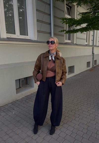 Chaqueta corta de cuero marrón, top marrón, pantalones de pierna ancha negros y botas de tobillo negras. Los accesorios incluyen una bufanda estampada y un bolso redondo pequeño.