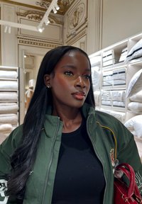 Mujer de cabello largo y negro, vestida con una chaqueta verde y sosteniendo una bolsa roja, está dentro de una tienda de ropa de cama de lujo con almohadas mullidas en los estantes.