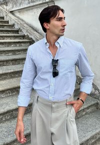 Camicia button-up a quadri blu chiaro con taschino sul petto, abbinata a pantaloni grigi chiari con pieghe. Occhiali da sole neri poggiati sulla camicia.
