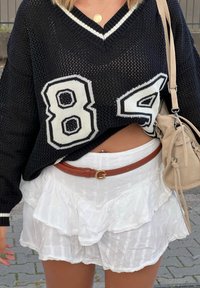 Μαύρος πλέγμα oversized πουλόβερ με άσπρες αριθμητικές λεπτομέρειες 84 και 5; σε συνδυασμό με λευκή υφασμάτινη φούστα με ρίγες και καφέ ζώνη. Ορατή είναι επίσης μια μπεζ τσάντα.