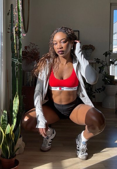 Mujer con sujetador deportivo rojo, pantalones cortos negros, chaqueta blanca y zapatillas, en cuclillas en una habitación iluminada por el sol con plantas de interior y objetos decorativos.