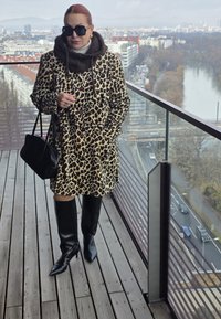 Manteau à imprimé léopard avec poches et col en fourrure marron, associé à des bottes noires à talons hautes jusqu'aux genoux. Sac à main noir, plancher en bois en dessous.