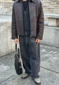 Dark brown leather jacket worn over a black ribbed sweater, teamed with loose-fitting black jeans. A black shoulder bag featuring silver hardware is carried in one hand.