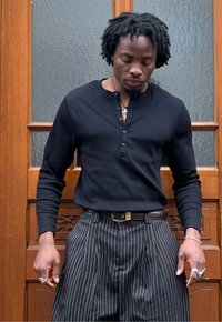 Camisa henley de manga longa preta com detalhe de botões e calças listradas. As mãos estão nos bolsos, exibindo um anel de prata e uma pulseira. Fundo de madeira.