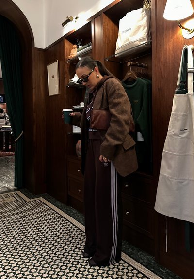 Blazer marrón texturizado sobre una sudadera oscura, combinado con pantalones de chándal burdeos con rayas blancas, sosteniendo una taza verde en un interior estiloso.