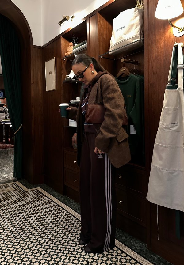 Blazer marrone testurizzato sopra una felpa scura, abbinato a pantaloni della tuta bordeaux con strisce bianche, mentre tiene in mano una tazza verde in un interno elegante.
