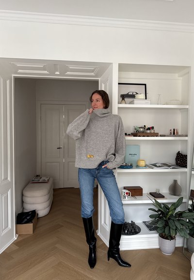 Mujer de pie en una habitación moderna, vestida con un suéter de cuello alto gris, jeans azules y botas negras hasta la rodilla, con estanterías y plantas cerca.