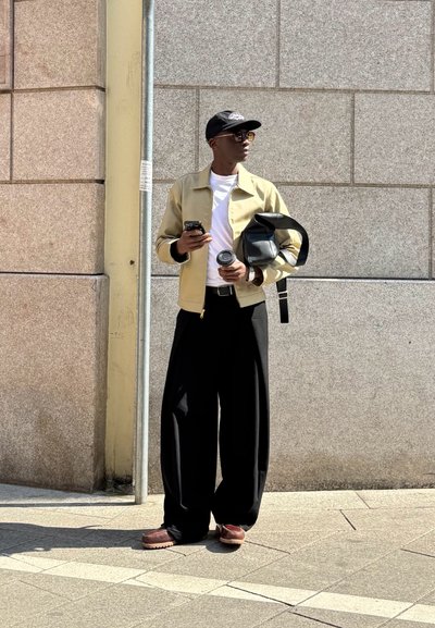 Una persona vestida con una chaqueta beige clara sobre una camisa blanca, pantalones anchos negros, sosteniendo una taza y un teléfono, de pie contra una pared gris.