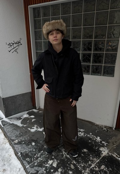 Chaqueta negra con dos bolsillos delanteros, pantalones marrones sueltos y un sombrero beige de piel. El fondo presenta una pared de vidrio texturizado y suelo nevado.