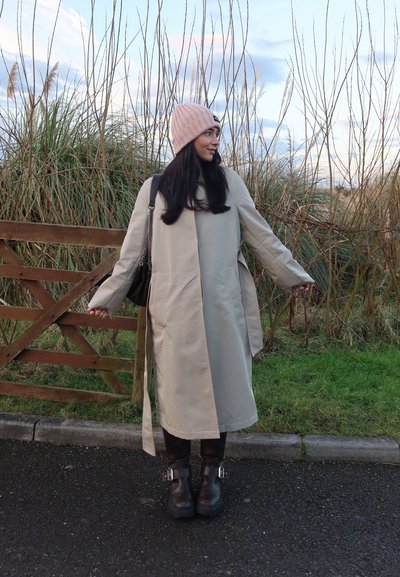 Abrigo largo color beige claro con aberturas laterales, usado sobre leggins negros y botas negras gruesas con hebillas decorativas. Gorra de lana rosa. Bolso de hombro marrón.
